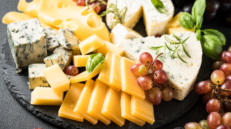 Variety of hard and soft cheeses with grapes and herbs on stone platter.