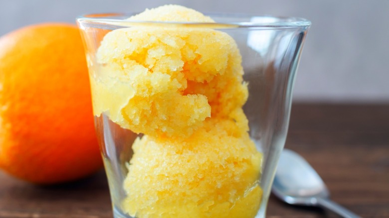 Two scoops of orange sherbet in small glass with whole orange and silver spoon in background