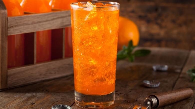 Glass of orange soda with ice on wooden table with wooden crate of orange soda bottles in background