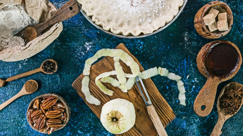 A peeled apple sits on a cutting board, surrounded by pie-makings: flour, sugar, warm spices, nuts, crust