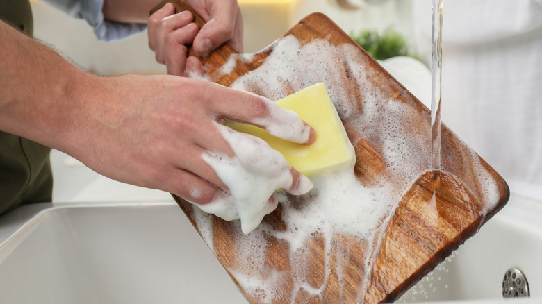person cleaning wooden cutting board with a sponge under running water