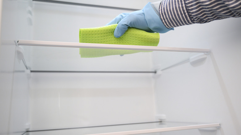 gloved hand wiping down the shelves of an empty fridge with a microfiber cleaning cloth