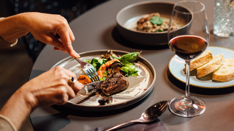 three plates on a table, meat and salad on one, a glass of wine, a plate of bread, and woman cutting meat