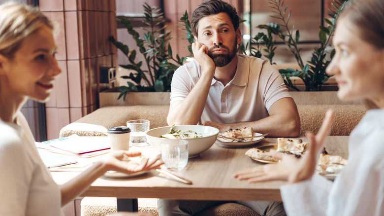 Bored-looking man sitting at a table in a restaurant watching two women talking in the foreground