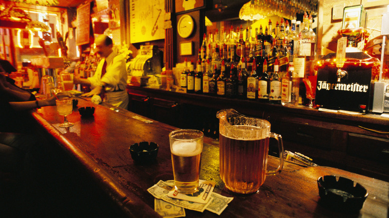 A shot down the bar at a dive bar with low, warm lights and a pitcher of beer and cash on the bartop