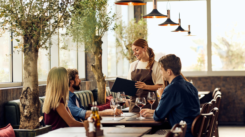 A group of friends being a waited on by a female server in a brown apron in a chic, modern restaurant