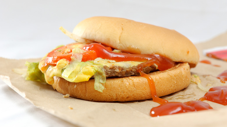 A messy looking hamburger with the top bun half off showing tomato, lettuce, and cheese on the patty. Ketchup is splattered on the burger and the paper next to it