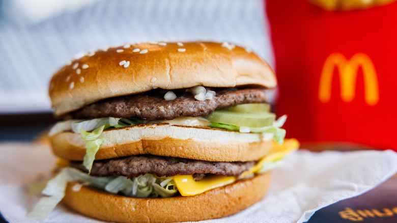 A Big Mac sitting on paper with the burger patties, pickles, lettuce, and cheese visible, and an out of focus box of fries in the background