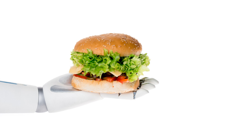 A robotic white hand, palm up, is holding a hamburger on a sesame seed bun that is loaded with lettuce, cheese, and tomato
