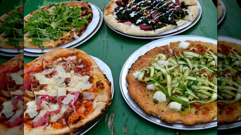 Assortment of four pizzas with various toppings, including cheese and greens, on a green wooden table