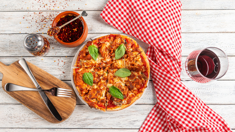 Wood-fired pizza with basil on a white wooden table with utensils, a glass of wine, jar of chili flakes, and red-and-white cloth