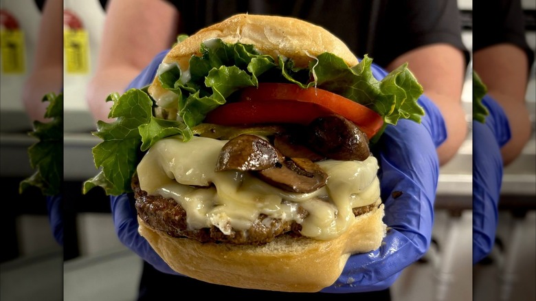 Hands holding a cheeseburger with mushrooms, lettuce, and tomato