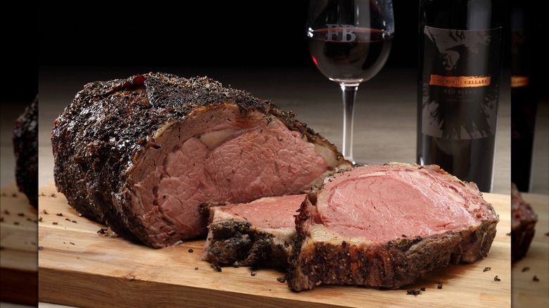 Whole roast and slices of prime rib on wood board with wine bottle and glass