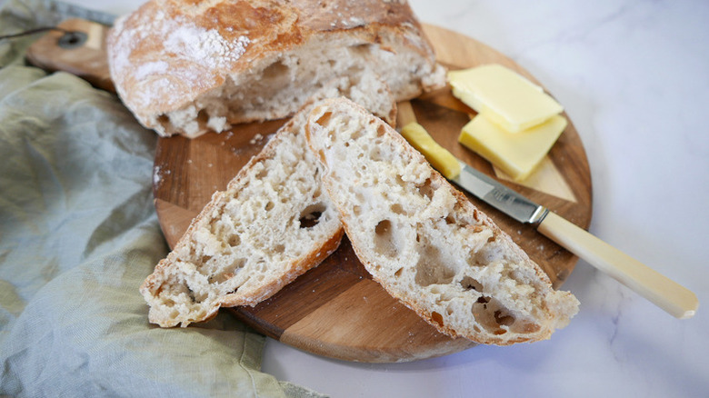 Sliced crusty loaf of bread on a wooden board with pats of butter and a knife