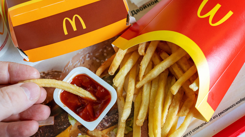 A hand dipping a McDonald's fry in ketchup with more fries nearby along with McDonalds branded packaging.
