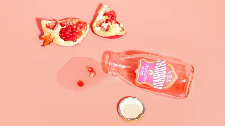 A bottle of spilled kombucha on a pink background laying beside a broken pomegranate