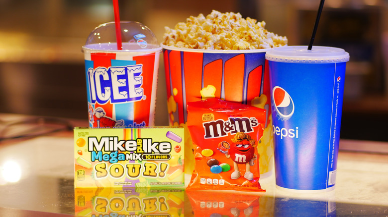 Old school ICEE, box of popcorn, soda cup, box of Mike and Ike's, and M&M's on a glass counter