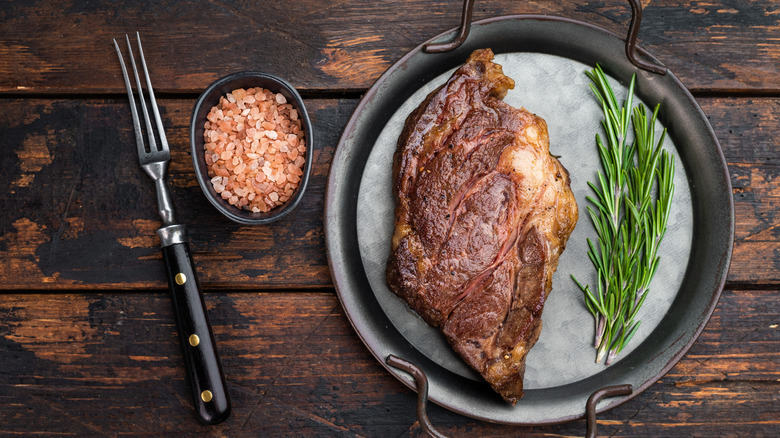 a seared steak on a metal tray with fresh rosemary, coarse salt, and a fork beside it atop a wooden table