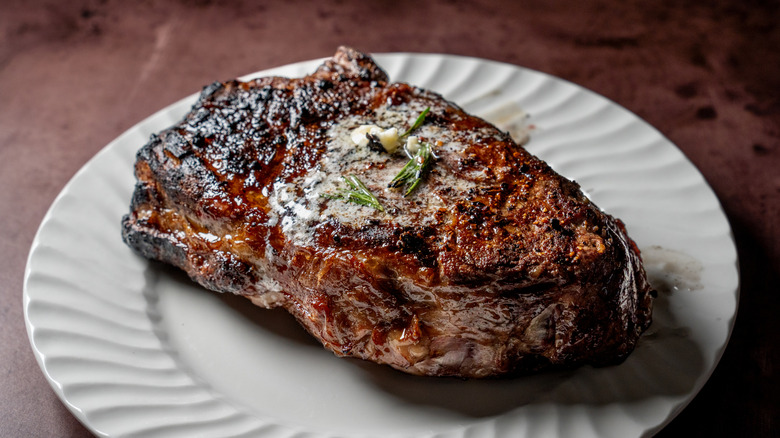 a seared ribeye topped with butter, garlic, and herbs on a white plate