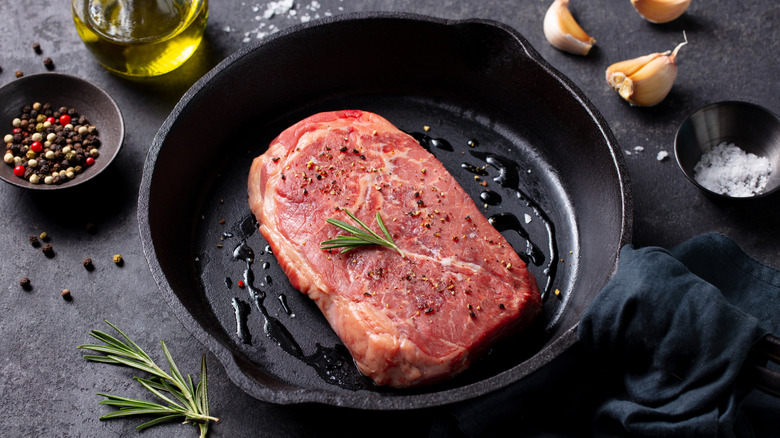 a raw steak in a cast iron pan with a drizzle of oil, surrounded by fresh herbs, garlic, spices, and a jar of olive oil