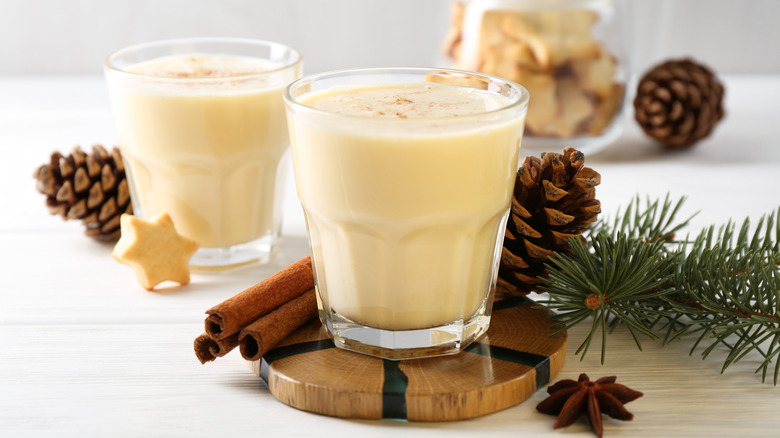 Two cups of eggnog surrounded by cinnamon sticks and pinecones.