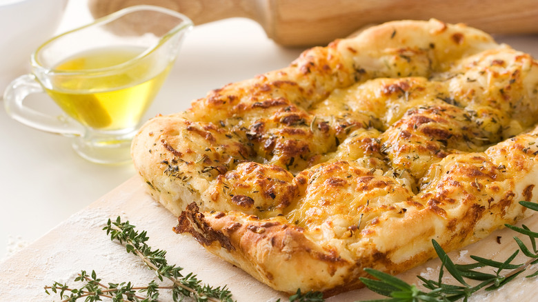 A loaf of bubbly focaccia next to olive oil and herbs