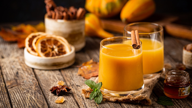 Two orange cocktails in short glasses surrounded by ingredients like mint, cinnamon, star anise, dried grapefruit slices, and gourds