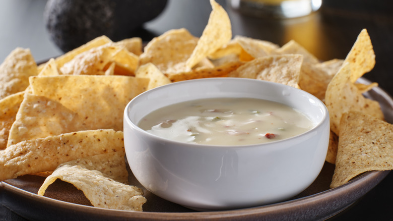 A small white bowl filled with queso dip on a plate with some tortilla chips