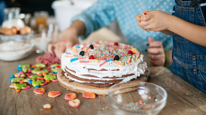 people decorating a layer cake with icing, sprinkles, and candy