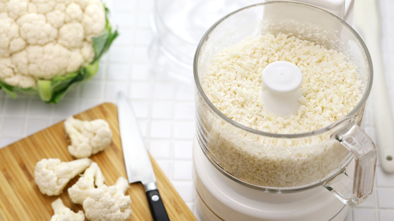 a full-sized food processor shown on a tiled countertop, with a whole cauliflower, florets on a cutting board next to a knife, and a work bowl filled with newly-crumbled cauliflower "rice"