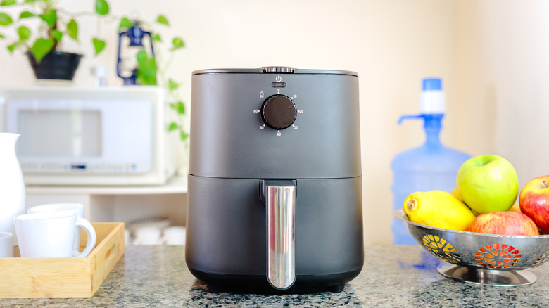 a sleek but simple air fryer in slate gray, shown on a kitchen counter between a fruit bowl and a tray containing a coffee/tea pot and cups