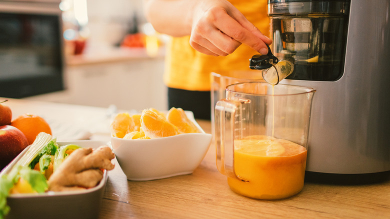 a young woman in sunshine-y yellow top and black pants holds open the spout on a juicer, which is filling a measuring cup with juice, next to bowls containing oranges, fresh ginger, celery, and other ingredients