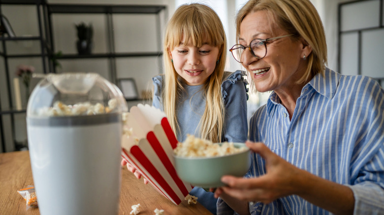 a mature woman and a young girl, both blonde-haired and dressed in blue, smiling and catching popcorn in their bowls as it hurtles from a hot air popper