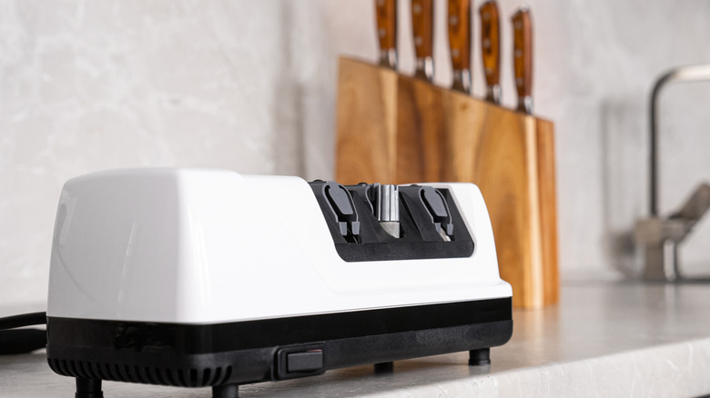 an electric knife sharpener with a black base and white body, pictured on a stone countertop next to a block filled with knives