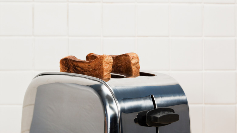 a toaster with a brushed-steel finish, shown against a white-tiled kitchen wall, with two pieces of well-toasted bread in its slots