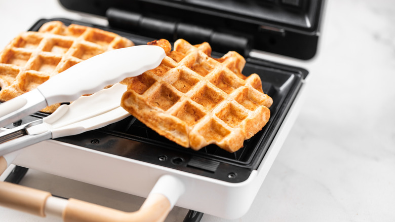 A pair of tongs with white silicone tips, removing the first of two waffles from a compact waffle maker