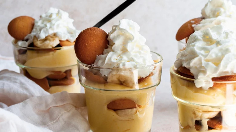 Three glasses full of banana pudding with vanilla wafers, banana slices, and whipped cream on top against pale background