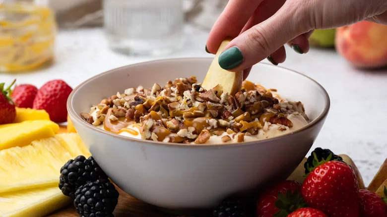 Hand dipping apple slice into white bowl of caramel and pecan dessert dip
