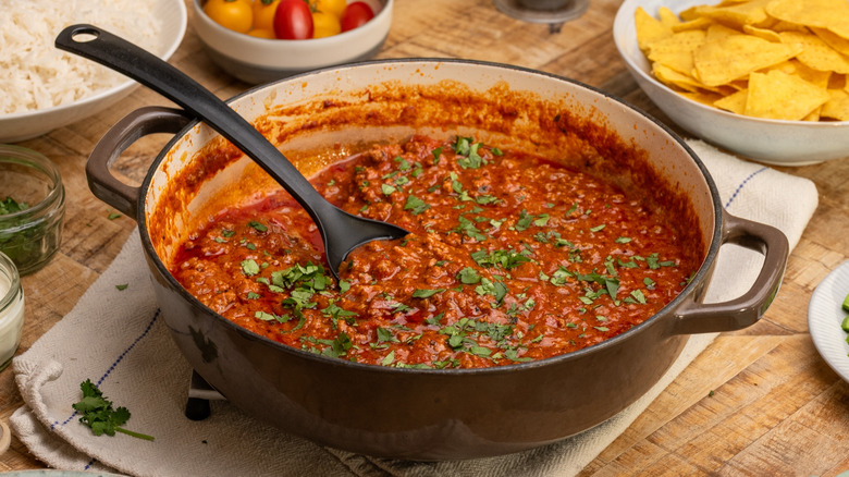 West Texas chili in pan with ladle surrounded by rice, cherry tomatoes, and tortilla chips