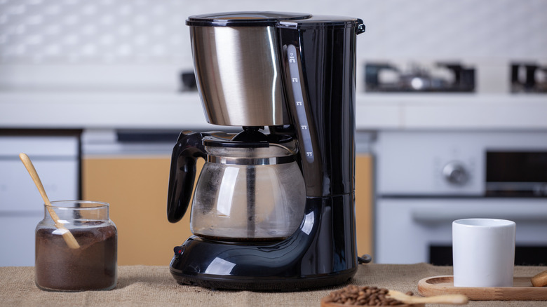 auto drip electric coffee maker with coffee grounds in a jar and a mug besides