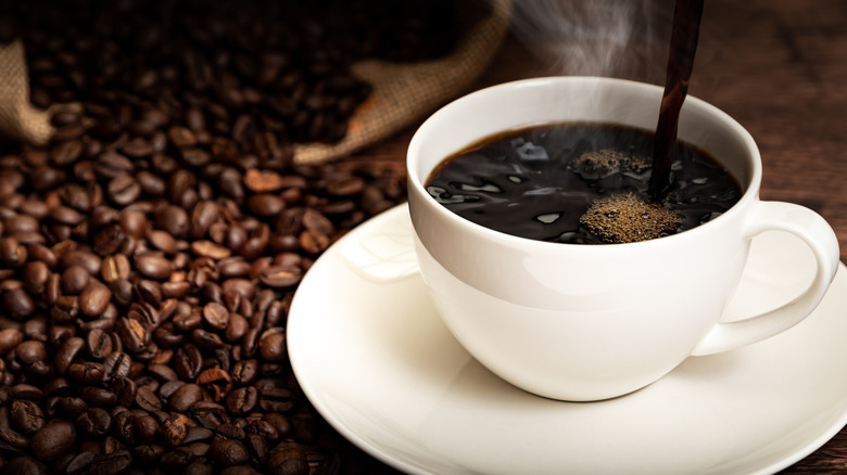 A cup of black coffee being poured surrounded by coffee beans