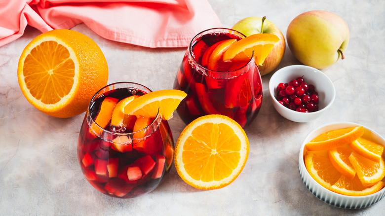 Two glasses of sangria on a table alongside some apples, sliced orange, and currants in a bowl