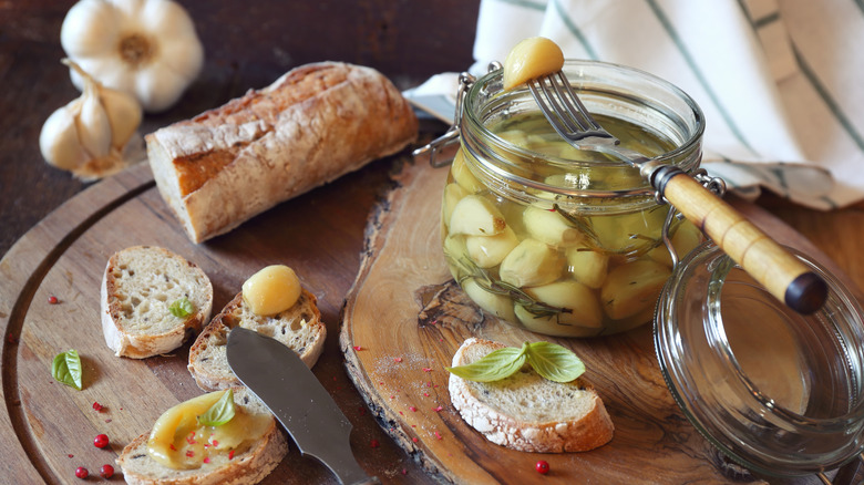 an open jar of garlic confit on a wooden board with bread and one clove skewer on a fork