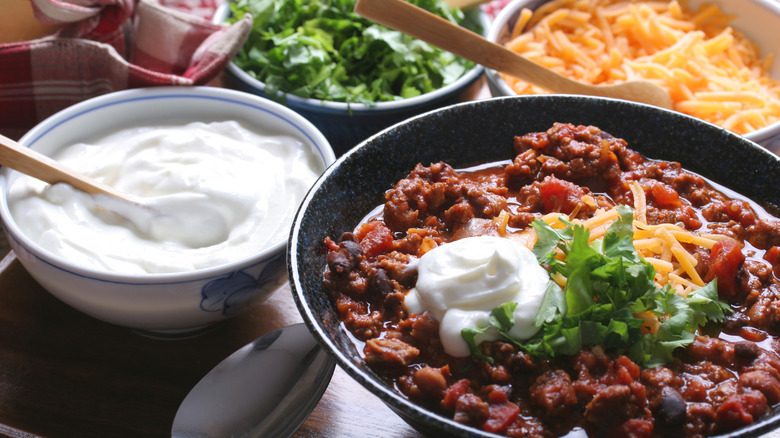 A bowl of chili surrounded by bowls of grated cheese, cilantro, and sour cream