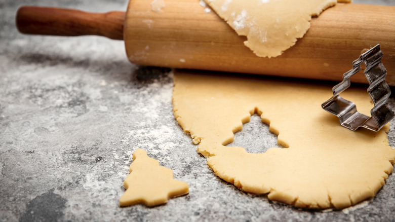 A rolling pin resting on some rolled cookie dough in a single Christmas tree cookie cut out with the cookie cutter balance next to it