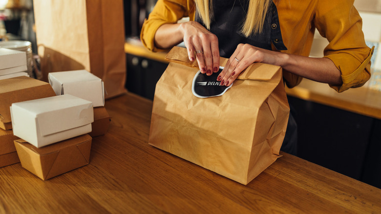 A person in a yellow shirt and apron sealing a paper bag for delivery with some to-go boxes on a wooden table in front of them