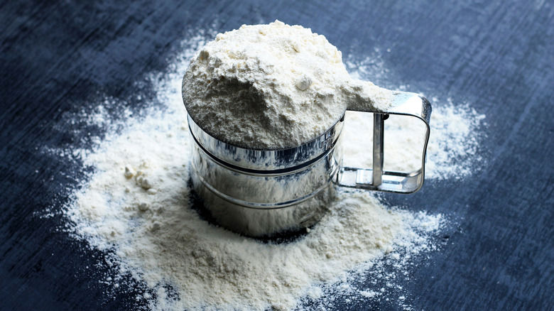 A sifter full of flour sits on a navy blue tablecloth with some flour spilled around it.