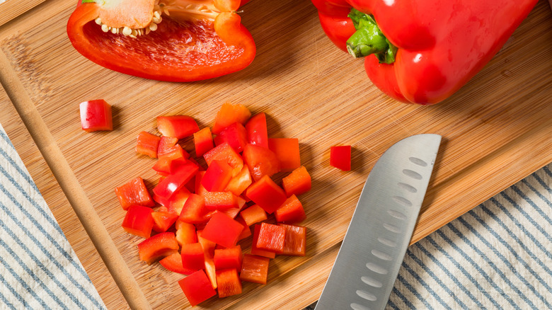 Half a red pepper and a full red pepper next to some chunks of sliced red pepper on a wooden cutting board with a knife